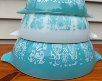May include: A stack of three nesting mixing bowls with side handles. The top and bottom bowls are turquoise with white graphic illustrations of people and produce. The middle bowl is white with turquoise graphic illustrations of the same pattern.