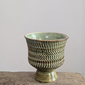 May include: A handcrafted ceramic goblet with a textured, woven-like pattern in shades of green and brown. The goblet has a wide, flared rim and a pedestal base, showcasing a rustic aesthetic. The interior is a lighter green.