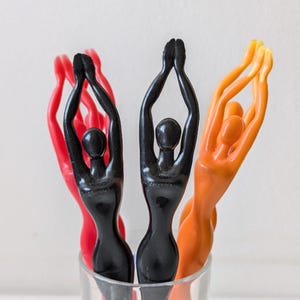 Könnte beinhalten: Vier dekorative Flaschenverschlüsse in einem Glas. Zwei sind schwarz, einer ist rot und einer ist orange. Die Verschlüsse sind in Form von stilisierten Figuren mit erhobenen Armen.