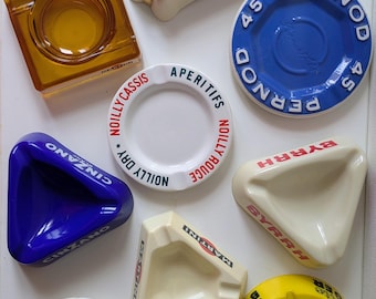 Puede incluir: Una colección de ocho ceniceros vintage de cerámica y vidrio con varios logotipos de marcas, incluidos Martini, Pernod 45, Noilly Prat, Cinzano, Byrrh, Suze y Berger. Los ceniceros varían en forma, incluyendo diseños triangulares, cuadrados y circulares.