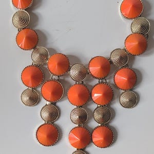 Könnte beinhalten: Eine auffällige Halskette mit goldfarbener Kette und Anhängern. Die Halskette hat abwechselnd orangefarbene, facettierte, runde Anhänger und strukturierte, goldfarbene, runde Anhänger. Ein auffälliges Schmuckstück.