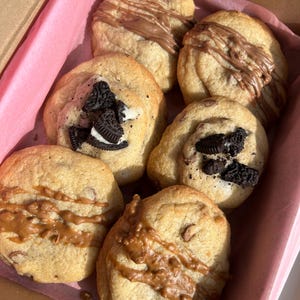 May include: A box containing six assorted cookies. The cookies are all round and have a light brown colour. Some of the cookies have chocolate chips, while others have chocolate or caramel drizzled on top. Some cookies have Oreo cookie pieces on top.