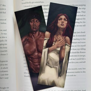 Puede incluir: Dos marcapáginas con ilustraciones de un hombre sin camisa con cabello oscuro y una mujer con cabello largo y rojo. El hombre se representa con una expresión seria, mientras que la mujer tiene una mirada contemplativa. Ambos marcapáginas tienen un aspecto pintado y texturizado.