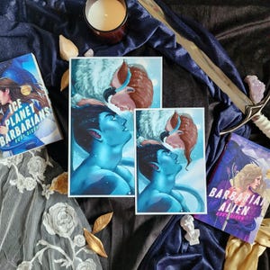 Puede incluir: Dos libros de bolsillo titulados Ice Planet Barbarians y Barbarian Alien de Ruby Dixon, junto a dos láminas rectangulares con un personaje de piel azul y una persona de pelo castaño, sobre tela azul oscuro con una espada de metal.