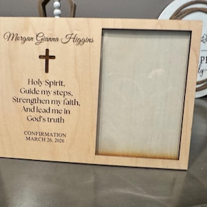 May include: A light-colored wooden photo frame with a rectangular opening. The frame is engraved with the name "Morgan Gianna Higgins", a cross, and a religious verse. The date "MARCH 26, 2026" is also engraved.