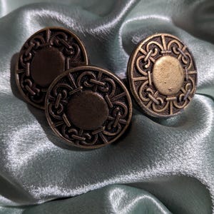 May include: Three antique bronze cabinet knobs with a circular design. Each knob features a central circle surrounded by an ornate, repeating pattern. The knobs are displayed on a light blue, silky fabric.