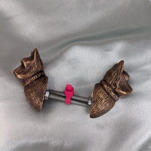 May include: A shoe stretcher with two bronze-colored dog head ornaments. The heads are connected by a metal rod, spring, and a pink plastic piece. The dog heads have detailed fur and collar designs.
