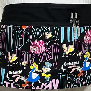 May include: Black waist apron with a colorful Alice in Wonderland design. The apron features Alice, the Mad Hatter, the Cheshire Cat, and the White Rabbit. Text includes "That Way", "This Way", and "Go back!". Two pens are in the pocket.