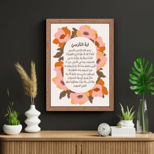 Könnte beinhalten: Gerahmter Druck mit arabischer Kalligraphie, umgeben von pfirsich- und rosafarbenen Blumenmustern. Das Kunstwerk wird auf einer Holzoberfläche mit dekorativen Pflanzen und einer Uhr präsentiert.