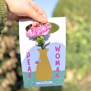 Könnte beinhalten: Eine weiße Karte mit einer Illustration einer rosa Nelke in einer gelben Vase. Auf der Karte steht in bunten Buchstaben "DEAR WOMAN". Der Hintergrund ist verschwommen grün.