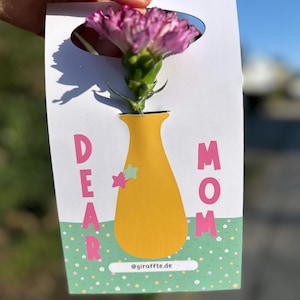 Könnte beinhalten: Eine weiße Karte mit einem Ausschnitt für eine rosa Blume, mit einem gelben Vasendesign und den Worten "DEAR MOM" in Pink. Die Karte hat einen grün-weißen Polka-Dot-Rand und die Website-Adresse @giraffte.de.