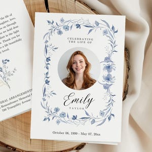 May include: A white memorial card featuring a photo of a woman, encircled by a blue floral wreath. The card says "Celebrating the Life of Emily Taylor" with dates below. Additional text is visible on a separate card.