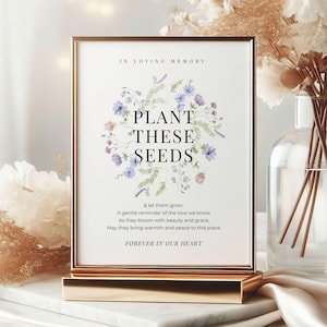 May include: A framed sign with the text "PLANT THESE SEEDS" surrounded by a floral wreath in shades of blue, purple, and green. The sign is in a gold frame and stand, with additional text below. The words "IN LOVING MEMORY" and "FOREVER IN OUR HEART" are also visible.