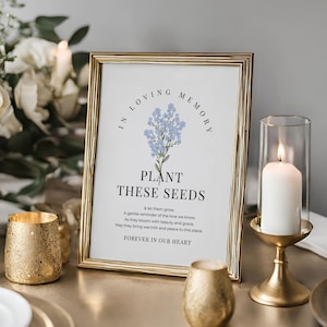 May include: A framed print with gold trim, featuring the text "IN LOVING MEMORY PLANT THESE SEEDS FOREVER IN OUR HEART." The print includes a blue floral illustration. A lit candle in a glass holder and gold candle holders are in the foreground.