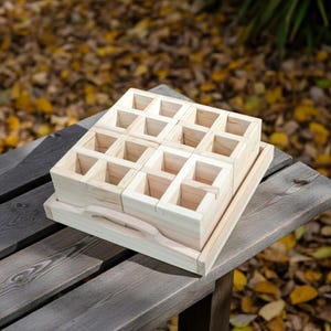 May include: A light-colored wooden organizer with multiple square compartments, stacked on a tray with a handle. The organizer is made of unfinished wood and sits on a wooden surface, with a blurred background of fall foliage.