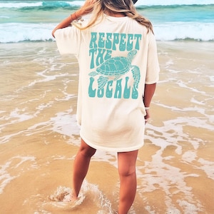 May include: A white t-shirt with a blue graphic of a sea turtle and the text "Respect the Locals" in a retro font.