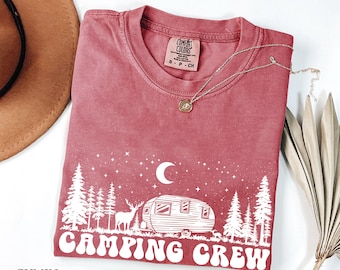 May include: A red t-shirt with a white graphic of a camper, trees, a deer, and a crescent moon. The text "Camping Crew" is written in a retro font.