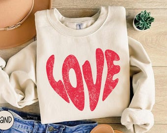 May include: A cream-colored sweatshirt with a red distressed graphic of the word "LOVE" in a heart shape.