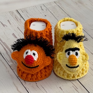 Op de afbeelding: Een paar handgemaakte gehaakte pantoffels, één oranje en één geel, elk met een gezicht van een personage met zwart haar, een grote neus en een lachende mond. De pantoffels zijn ontworpen om op populaire stripfiguren te lijken.