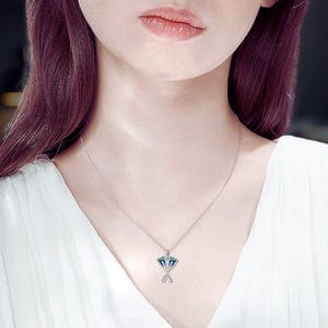 Puede incluir: Un collar plateado con un colgante con un diseño geométrico con detalles azules y verdes. El colgante está suspendido de una delicada cadena. La persona de la imagen lleva una blusa blanca.