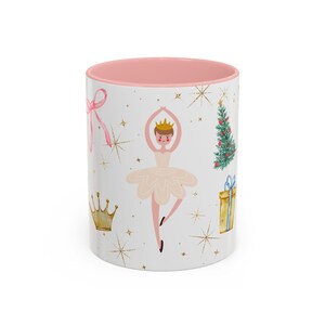 Puede incluir: Taza de cerámica blanca con interior rosa, decorada con una bailarina, un árbol de Navidad, un regalo, una corona e ilustraciones de lazo. Los estallidos de estrellas doradas y los acentos completan el diseño. La taza es de tamaño estándar.