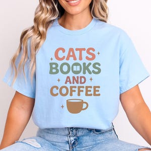 Puede incluir: Camiseta azul claro con las palabras "CATS BOOKS AND COFFEE" en una fuente retro. El texto es en tonos naranja, verde y marrón, con un dibujo de taza de café. La camiseta es de estilo casual.