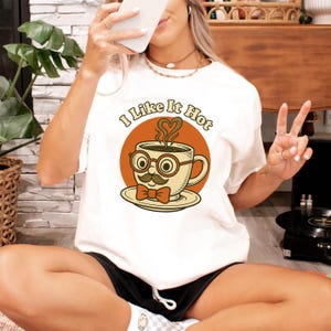 Op de afbeelding: Crèmekleurig T-shirt met een cartoon koffiekopje. De beker heeft een snor, bril en een strikje, met de tekst "I Like It Hot" erboven. De stoom uit de beker heeft de vorm van een hart, tegen een oranje cirkel.