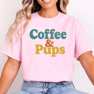 Puede incluir: Camiseta rosa claro con la frase "Coffee & Pups" en letras turquesa, naranja y amarillo. Esta camiseta informal está hecha de una tela suave, perfecta para el uso diario.