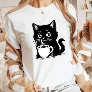 Può includere: T-shirt bianco con un disegno di un gatto nero dei cartoni animati che tiene in mano una tazza di caffè bianca da cui esce vapore. Il gatto ha grandi occhi e un aspetto soffice. Il design è un'illustrazione semplice in bianco e nero.