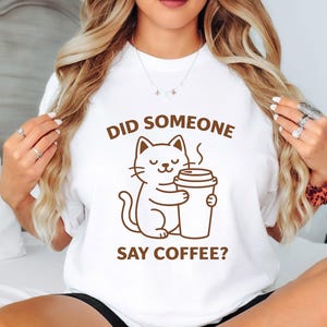 Peut inclure: T-shirt blanc avec un dessin marron d'un chat tenant une tasse de café. Le texte "DID SOMEONE SAY COFFEE?" est imprimé au-dessus et en dessous du chat.