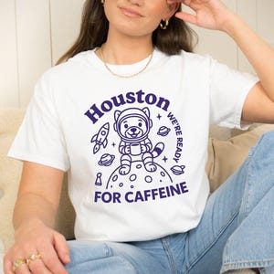 May include: White t-shirt with a purple graphic. It features a cartoon bear in an astronaut helmet holding a coffee cup. The text reads "Houston we're ready for caffeine" with a rocket and planets.
