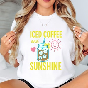 May include: White t-shirt with the text "ICED COFFEE and SUNSHINE" in yellow. The design includes a pink heart, a smiling sun, and a mason jar filled with ice cubes and coffee. A blue straw and lemon slice are also included.
