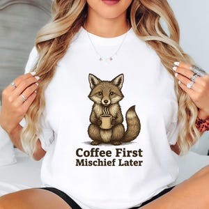 Könnte beinhalten: Weißes Sweatshirt mit einem Cartoon-Fuchs, der eine Kaffeetasse hält. Der Fuchs ist braun und beige. Der Text "Coffee First Mischief Later" ist darunter in brauner Schrift gedruckt.