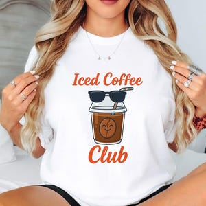 Op de afbeelding: Wit T-shirt met de woorden "Iced Coffee Club" in oranje. De afbeelding toont een cartoon ijscoffiebeker met een zonnebril en een lachend koffieboongezicht. Het shirt is een casual stijl.