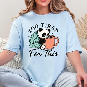 Può includere: T-shirt azzurra con un panda dei cartoni animati in una tazza color pesca, che indossa un pigiama. Il testo dice "TOO TIRED For This". La maglietta ha una vestibilità ampia. Il disegno è stampato sul davanti.