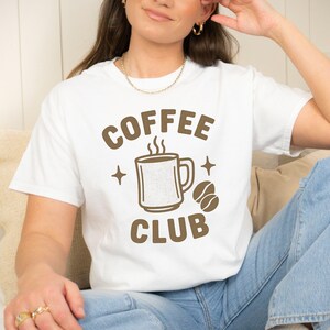May include: White t-shirt featuring a brown graphic with the words "COFFEE CLUB." The design includes a steaming coffee mug, coffee beans, and star accents. A casual, everyday wear item.