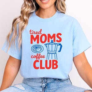 May include: Light blue t-shirt with the words "tired MOMS coffee CLUB" in red and blue. The design includes a coffee pot and a pastry illustration. A casual, comfortable tee.