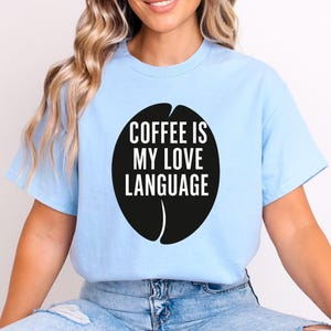 May include: Pale blue t-shirt featuring a black coffee bean design. The graphic includes the white text "COFFEE IS MY LOVE LANGUAGE". This casual, short-sleeved top is a relaxed fit.