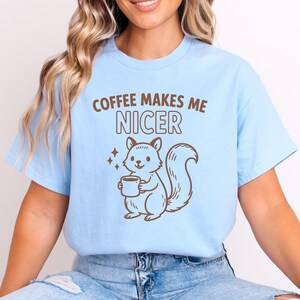 May include: Pale blue t-shirt featuring the phrase "COFFEE MAKES ME NICER" and a brown squirrel illustration holding a coffee cup. The squirrel has a bushy tail and is smiling. The shirt is a relaxed fit.