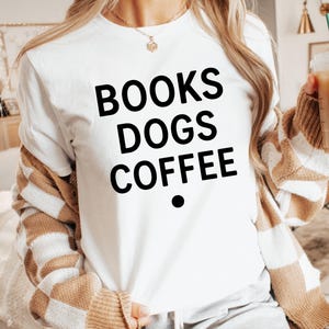 Puede incluir: Camiseta blanca con las palabras "BOOKS DOGS COFFEE" en letras negras grandes. La camiseta se lleva con una rebeca a rayas beige y blancas. Un punto negro está debajo de la palabra "COFFEE".