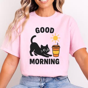 Peut inclure: T-shirt rose clair avec l'inscription "GOOD MORNING" en noir. Un chat noir s'étire vers une tasse de café et un soleil jaune. Le t-shirt est fait d'une matière douce.