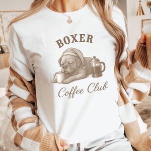 Puede incluir: Camiseta blanca con un gráfico de un perro bóxer con gorro de dormir, sosteniendo una taza de café y una cafetera francesa. El texto "Boxer Coffee Club" está impreso encima y debajo de la ilustración del perro. Prenda de vestir informal.