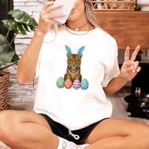 Può includere: T-shirt bianco con una tigre con orecchie da coniglio blu e uova di Pasqua colorate. La tigre ha strisce arancioni e nere. Le uova sono verdi, rosse, viola e blu con motivi. Un design festoso.