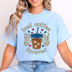 May include: Pale blue t-shirt featuring a graphic of an iced coffee in a clear cup with ice cubes, a blue lid, and a striped straw. The words "iced coffee forever" are in a circular design, with skeleton hands holding the cup. Red hearts and coffee beans are scattered around.