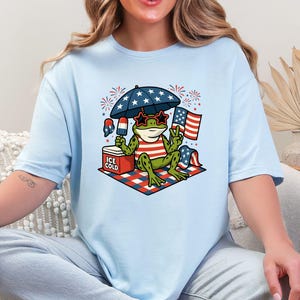May include: Light blue t-shirt featuring a cartoon frog wearing star-shaped sunglasses and a red, white, and blue striped shirt. The frog is under a star-covered umbrella, holding a popsicle, and sitting on a picnic blanket with an American flag.