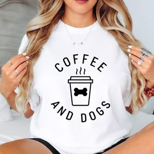 Puede incluir: Camiseta blanca con las palabras "COFFEE AND DOGS" en negro, rodeando un gráfico negro de una taza de café con un hueso de perro. La camiseta es de estilo casual.