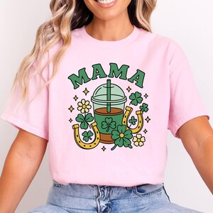 May include: A light pink t-shirt with the word "MAMA" in green block letters. The shirt features a graphic of a coffee drink, shamrocks, horseshoes, and flowers in shades of green, yellow, and brown.