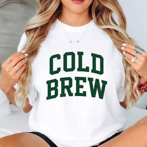 Puede incluir: Camiseta blanca con la frase "COLD BREW" en letras verdes oscuras y en negrita. La camiseta es de estilo casual, adecuada para el uso diario. El texto está centrado en la parte delantera.