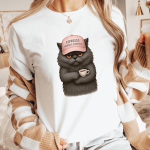 Op de afbeelding: Wit T-shirt met een afbeelding van een chagrijnige grijze kat met een roze pet met de tekst "Espresso Yourself, Carefully". De kat houdt een kopje koffie vast. Het shirt is een casual stijl.