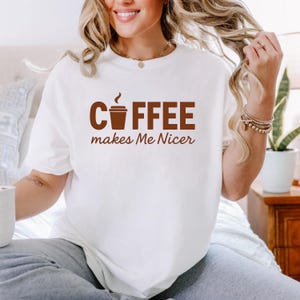 Puede incluir: Camiseta blanca con el texto marrón "COFFEE makes Me Nicer" y un gráfico de taza de café. La camiseta es de estilo casual, adecuada para el uso diario.